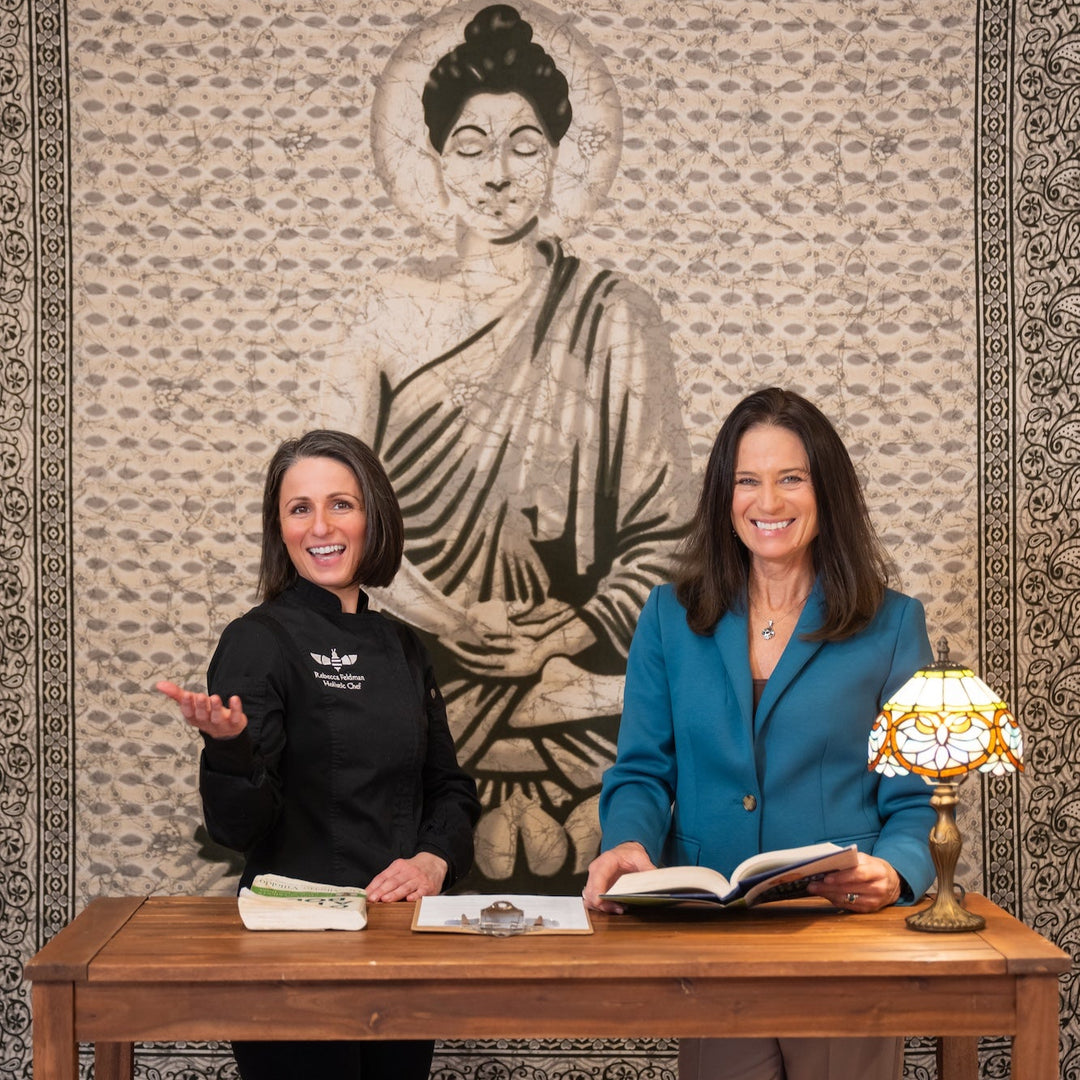 Two women standing behind a table with books, smiling in front of a tapestry with Buddha's image.