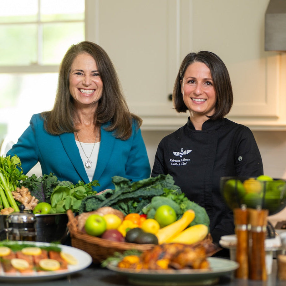 Two women in a kitchen with a variety of fruits and vegetables on the counter.