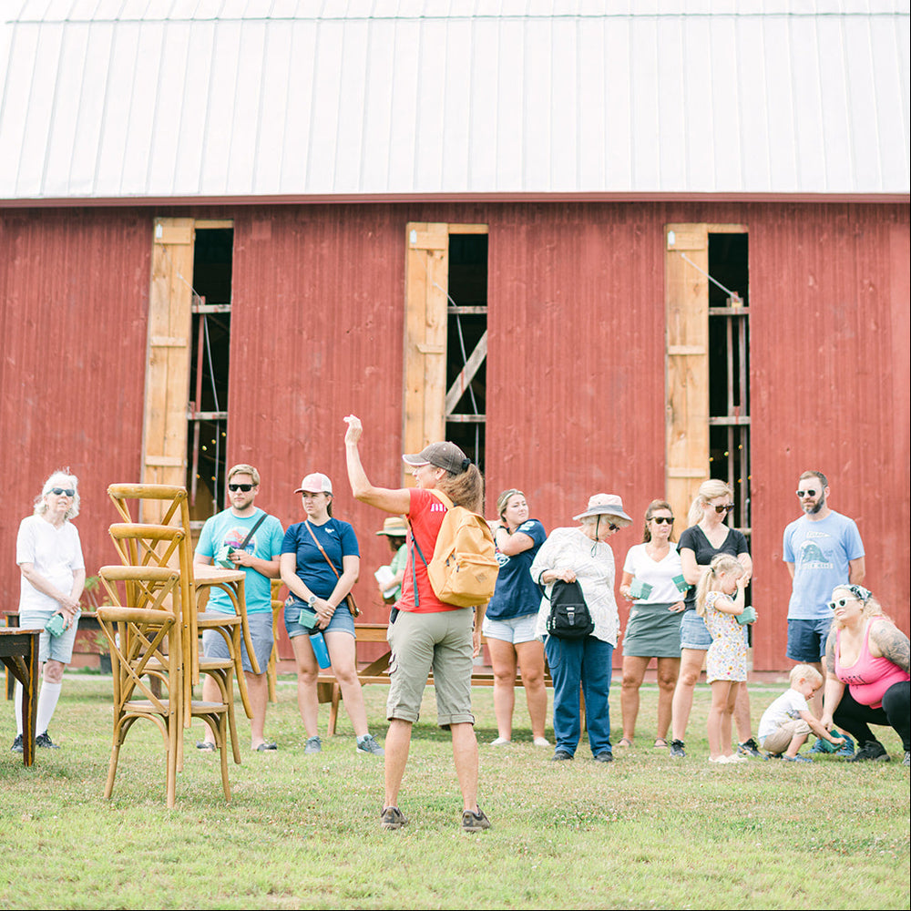 A group of people standing in front of a red barn on a grassy field, with one person in the center raising their arm. The scene appears to be outdoors during a tour or event, with the barn and open sky visible.
