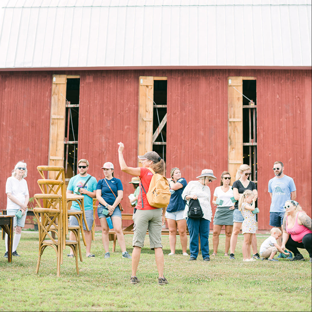 A group of people standing in front of a red barn on a grassy field, with one person in the center raising their arm. The scene appears to be outdoors during a tour or event, with the barn and open sky visible.