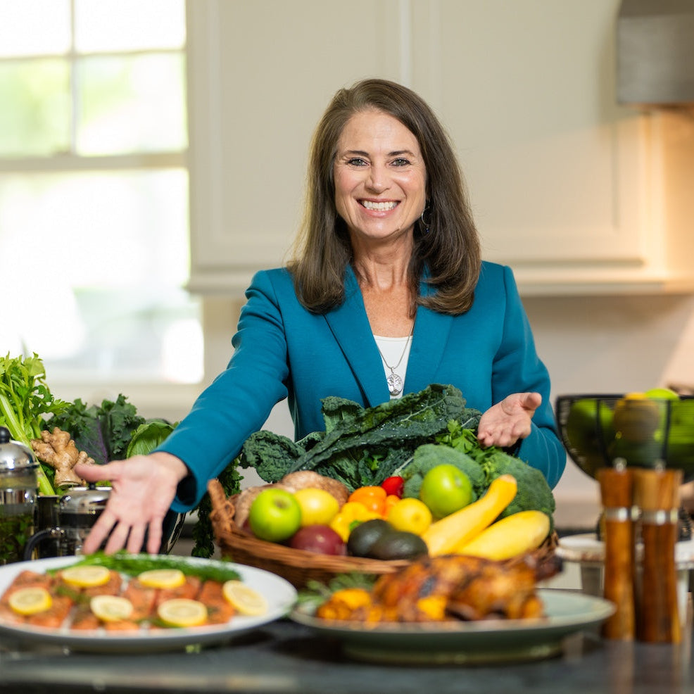 A smiling woman in a teal blazer stands in a bright kitchen behind a counter filled with fresh vegetables, fruits, and prepared dishes, gesturing toward the food.