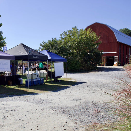 Outdoor event scene with several pop-up canopy tents set up on a gravel driveway in front of trees and a red barn. People are gathered around tables under the tents, and a large white banner is visible on one tent.