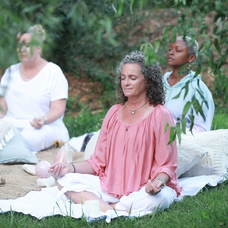 Three women sitting on a blanket outdoors, with one woman meditating in the foreground.