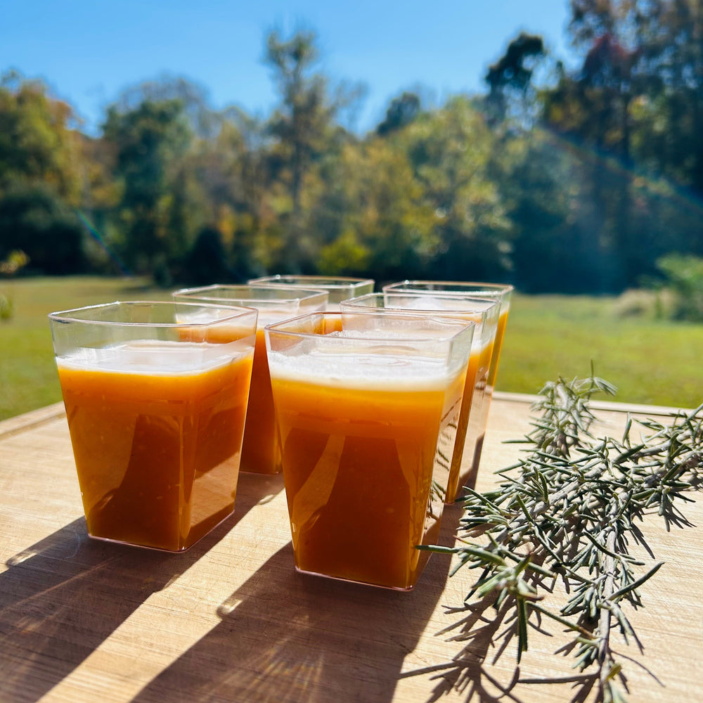 Clear cups of an orange liquid on a wooden cutting board with some sprigs of rosemary. There are blurred fall trees and blue sky in the background.