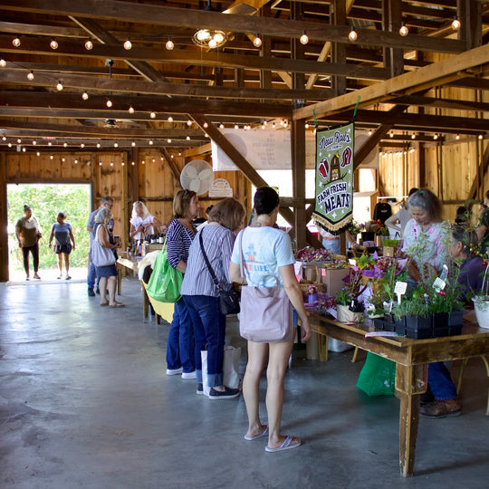 A bustling indoor farmers market scene inside a rustic barn with exposed wooden beams and string lights. Several people are standing at tables displaying flowers, plants, and produce, with a sign reading 'Local Farm Fresh' hanging on the right.