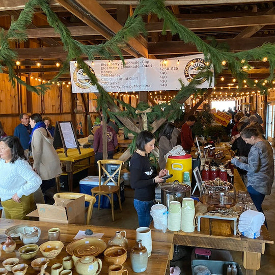A bustling indoor market or craft fair held in a rustic wooden barn, with people gathered around tables displaying handmade pottery, mugs, cups, and other home goods. Green garlands and warm string lights hang overhead, creating a cozy, rustic atmosphere.