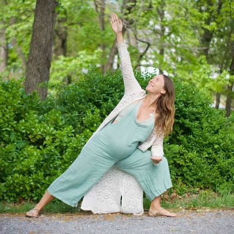 Pregnant woman in green overalls and a white cardigan doing yoga in a park.