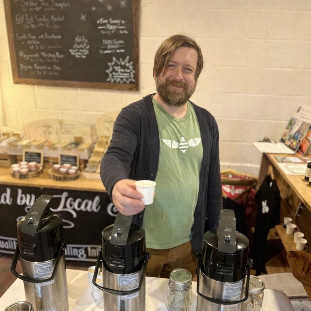 Man holding a white cup next to stainless steel canisters on a table with a chalkboard menu in the background.