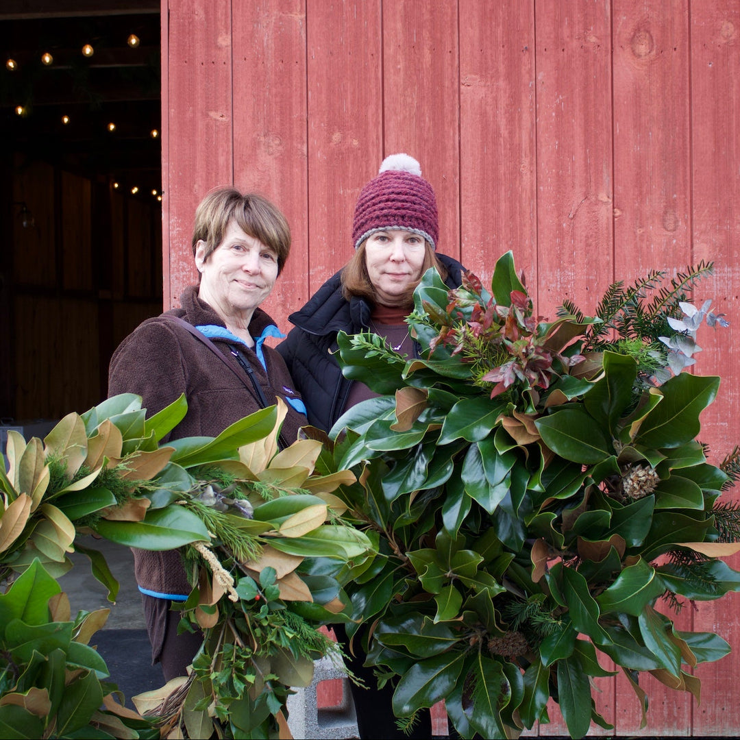 Two women standing in front of a red barn holding large holiday wreaths made of green leaves and evergreen branches.