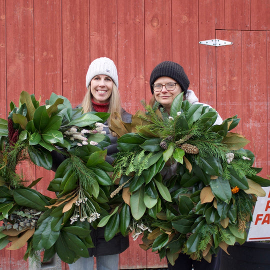 Two women standing in front of a red barn holding large green holiday wreaths.