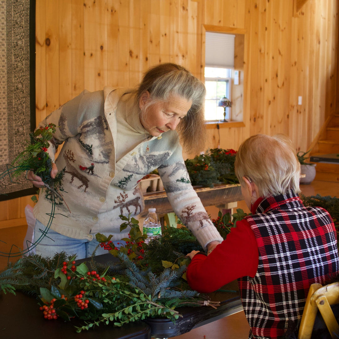Two people gathered indoors around a table, working on floral arrangements with greenery and red berries. The room has wood-paneled walls and a large framed artwork on the left.