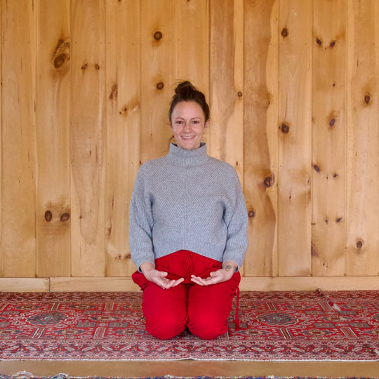 Person in gray sweater and red pants sitting on a rug in front of a wooden wall.