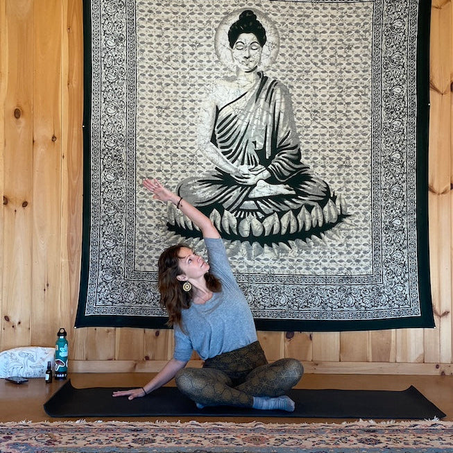 Person practicing yoga in front of a tapestry with a Buddha design on a wooden wall.