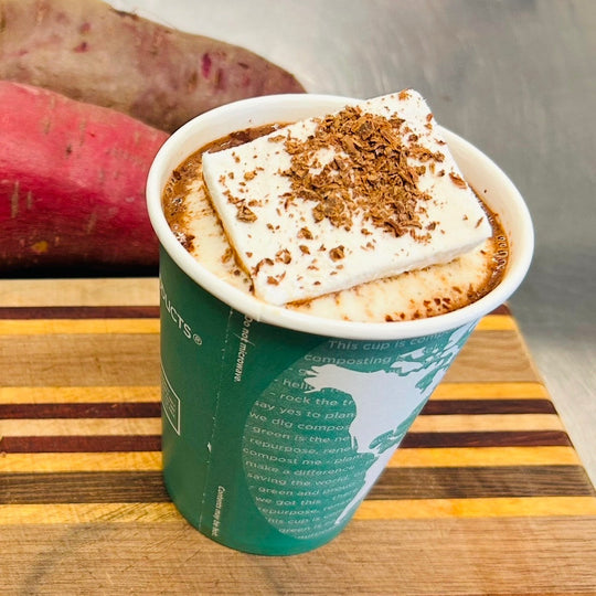 Green paper cup of hot chocolate with marshmallow and shaved chocolate sitting on a wooden cutting board with Japanese sweet potatoes in the background.