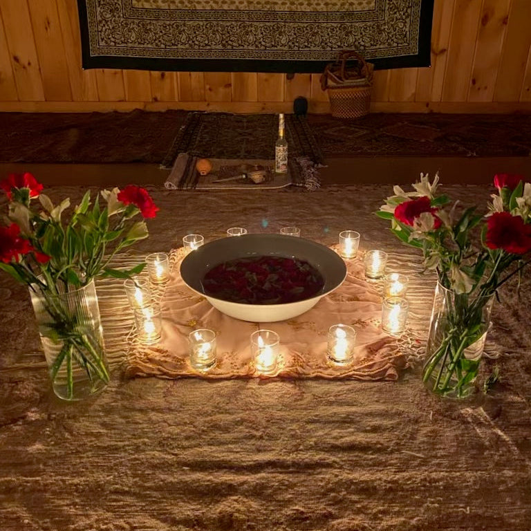 A ritual or meditation setup on a rustic wooden table, featuring a central white bowl filled with dark red powder, surrounded by numerous lit tealight candles. Two glass vases filled with red and white flowers sit on either side.