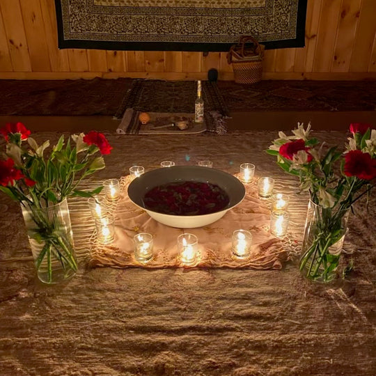 A ritual or meditation setup on a rustic wooden table, featuring a central white bowl filled with dark red powder, surrounded by numerous lit tealight candles. Two glass vases filled with red and white flowers sit on either side.