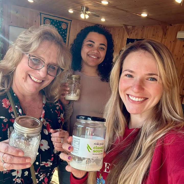 Three smiling women in a wooden space holding jars of sourdough starter
