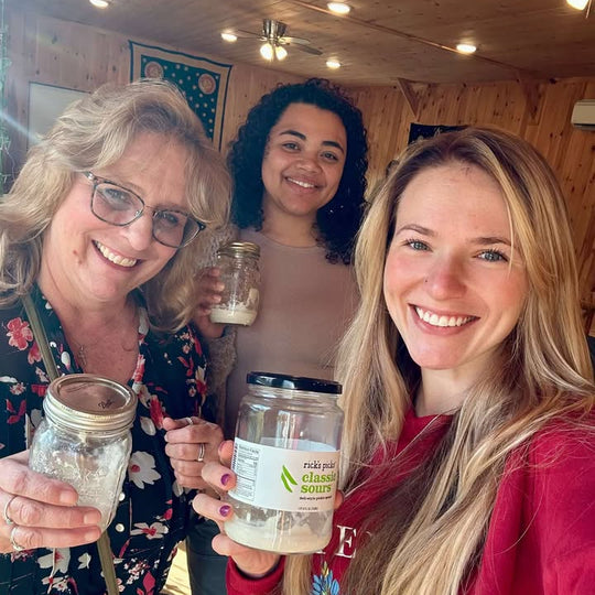 Three smiling women in a wooden space holding jars of sourdough starter