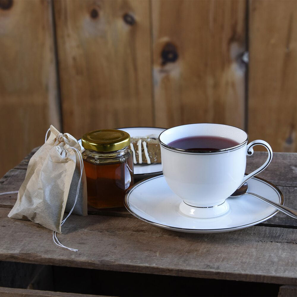 A cup of tea on a saucer with a tea bag on the side, next to a jar of honey, all placed on a wooden surface.