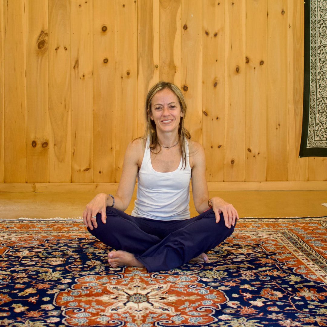 Woman sitting in a yoga pose on a patterned rug with wooden walls.