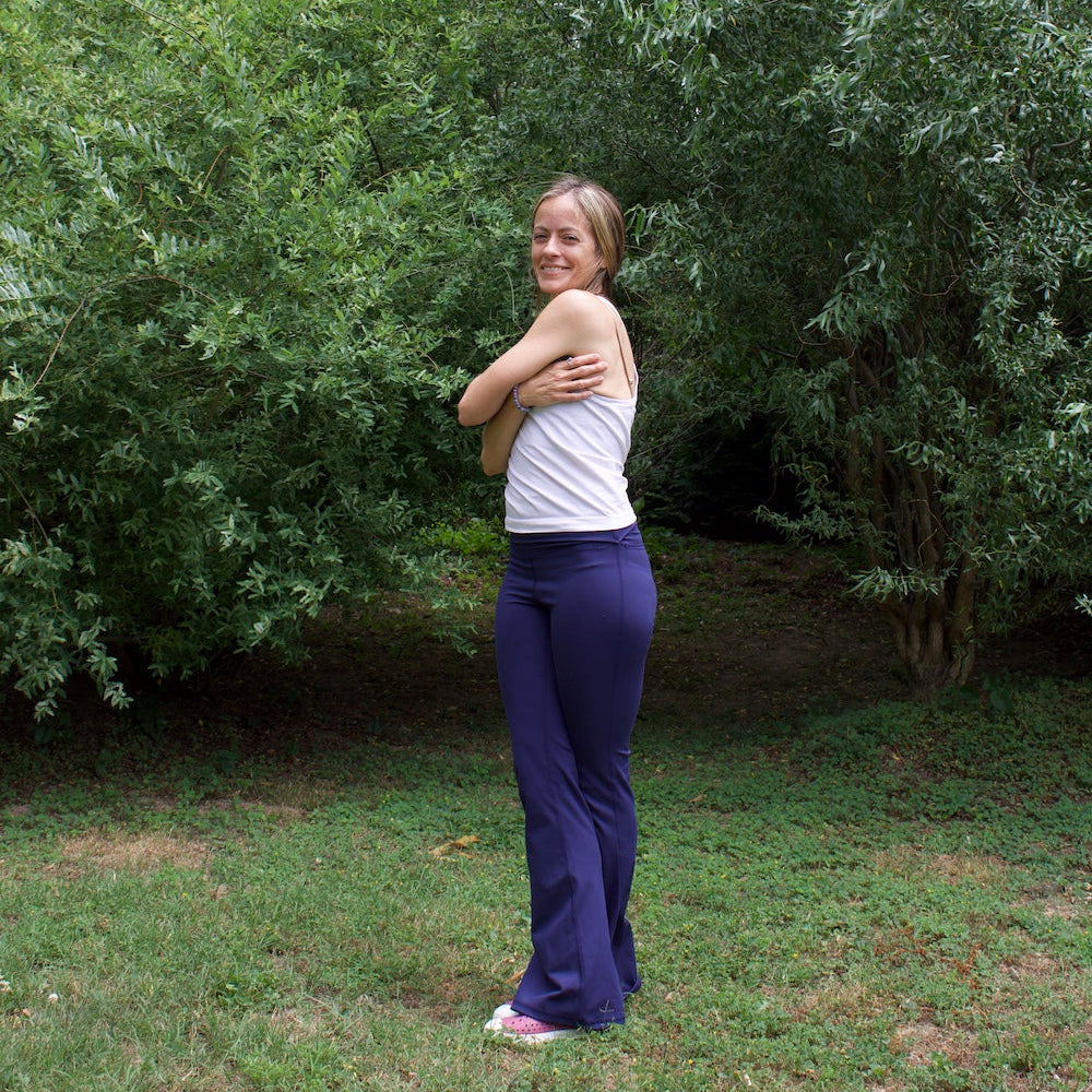 Woman in a white tank top and blue yoga pants doing a twist in front of green shrubbery