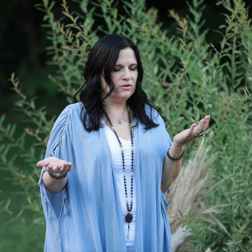 Woman in a blue robe standing outdoors with greenery in the background