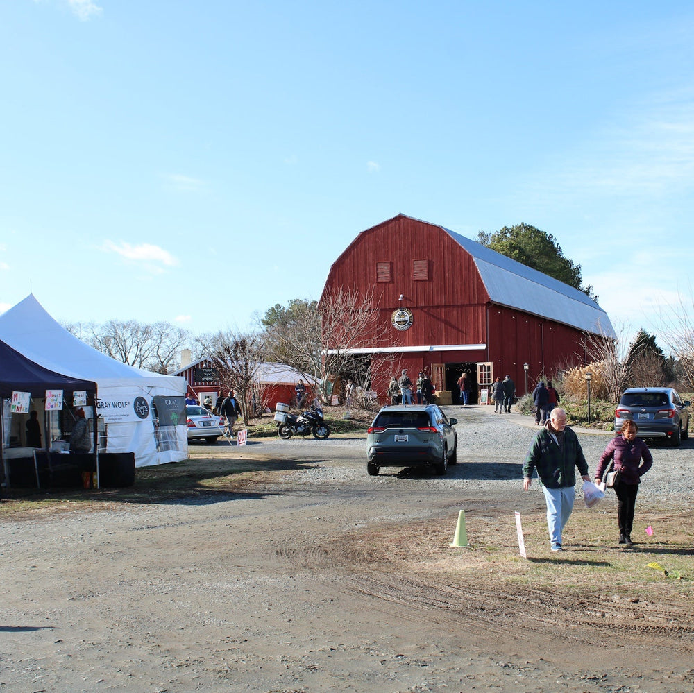 A large red barn with a white roof stands in the background of an outdoor farm event, with tents on the left, parked cars on the right, and a few people walking in the foreground on a dirt lot under a blue sky.