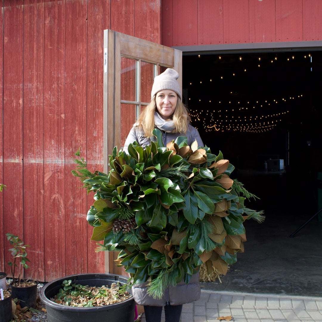 Person holding a large green wreath in front of a red barn with open doors.
