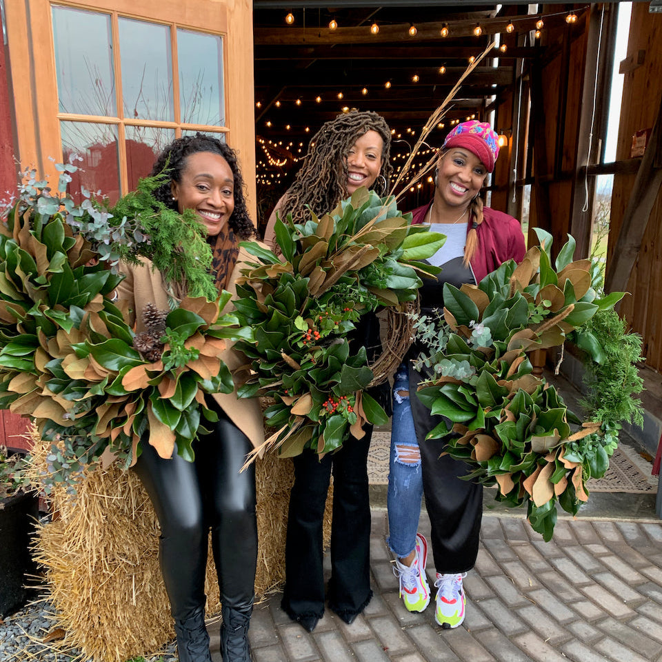 Three women holding large green wreaths in front of a barn.